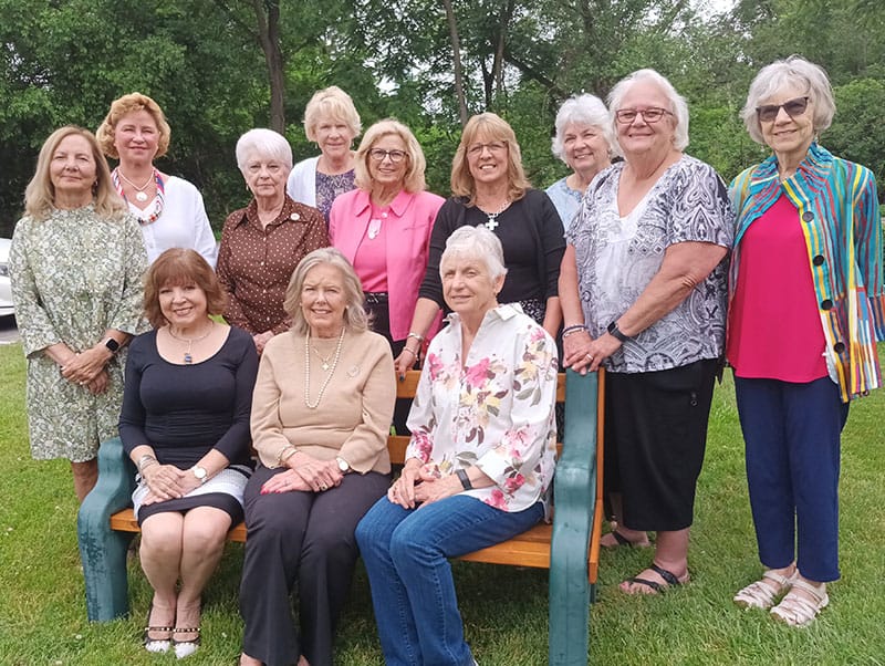 Group of older women posing together in a park, standing and sitting around a wooden bench on a sunny day.