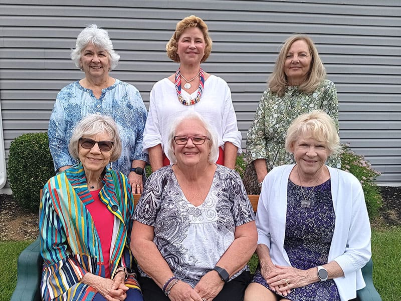 Womans-Club-of-York-2026-27-Board Six smiling women posing outdoors in front of a gray siding building, seated and standing in two rows.